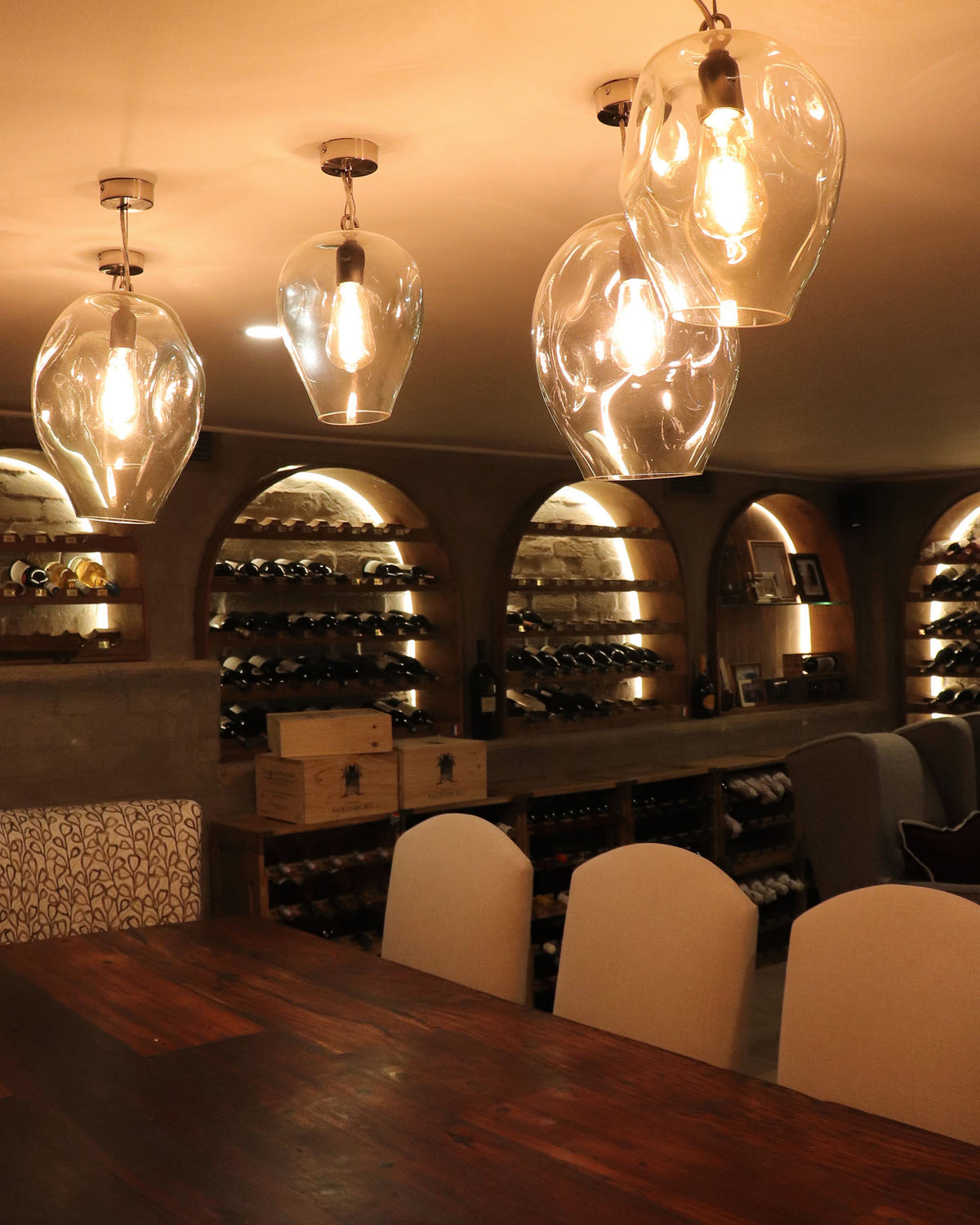A set of modern orb glass pendant light fixtures over a custom timber dining table in a warmly lit wine cellar or restaurant setting.